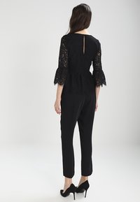 Černý krajkový top s rozšířenými rukávy a peplum pasem, v kombinaci s úzkými černými kalhotami. Výrazný otevřený klíčový otvor na zádech a strukturovaný vzor.