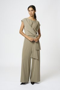 Beige jumpsuit with a draped design, short sleeves, and a belted waist, featuring wide legs and a smooth fabric texture. Black pointed-toe heels.