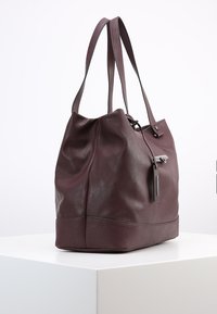 Dark burgundy leather tote bag with a soft texture, two shoulder straps, and a side zipper detail, featuring a flat bottom for stability.