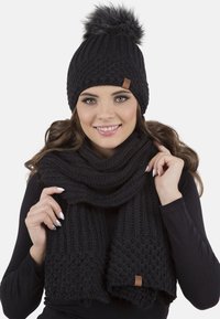 Black knitted beanie with a faux fur pom-pom, paired with a matching thick knitted scarf, both featuring a textured design and leather tag.