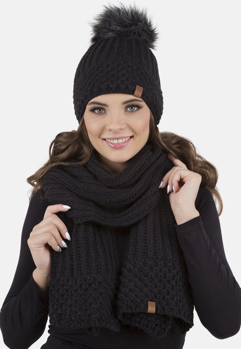 Sort strikket hue med en faux fur pom-pom, parret med et matchende tykt strikket tørklæde, begge med et struktureret design og lædermærke.