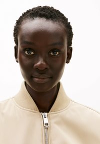 Giacca bomber beige con colletto a coste, zip frontale e tessuto liscio. Il modello ha i capelli corti e scuri e un'espressione neutra.