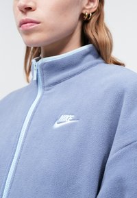 Svetlo modrá mikina na zips z flísu s vysokým golierom, ktorá má biele logo Nike na hrudi a kontrastné modré zipsy.