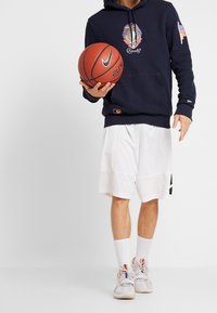 Mörkblå hoodie med broderad logotyp och amerikansk flagga, ihop med vita basketshorts och grå sneakers med multicolorerade detaljer, hållande en basketball.
