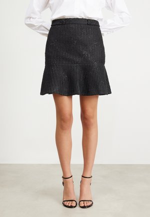 Femme portant une mini-jupe noire texturée avec un ourlet évasé, assortie à des talons noirs à brides à la cheville et une chemise blanche à manches longues.