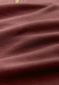 Bordo spalvos audinys su lygiu, tekstūruotu paviršiumi, kuriame yra subtilus mezgimo raštas. Medžiaga atrodo minkšta ir natūraliai krenta.