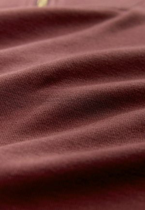 Burgundowa tkanina o gładkim, fakturowanym wykończeniu, z subtelnym wzorem dzianiny. Materiał wydaje się miękki i naturalnie się układa.