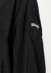 Jachetă neagră cu o suprafață texturată, prezentând un patch pe umărul stâng cu logo-ul "Blauer." în gri pe un fundal negru.