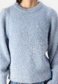 Femme portant un pull en tricot flou bleu clair avec des fils métalliques, associé à un jean en denim bleu et de grandes boucles d'oreilles.