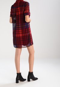 Abito camicia a quadri rosso e blu con maniche corte, dettagli con bottoni e una texture traslucida, abbinato a stivali neri alla caviglia e una silhouette moderna.