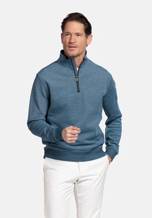 Man draagt een blauwe trui met een rits tot aan de borst en witte broek, staat met één hand in de zak en de andere licht verheven.