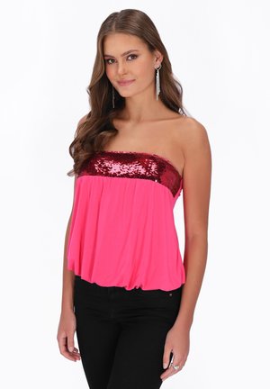 Jonge vrouw die een strapless roze top met rode pailletten en zwarte broek draagt, glimlachend met lang golvend bruin haar en hangende hartvormige oorbellen.