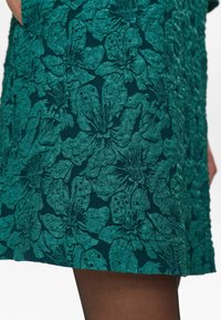 Rochie teal cu un model floral texturat, având petale verzi ridicate și accente închise. Țesătura este elastică, cu o finisare netedă.