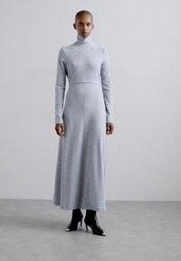 Grijze jurk met lange mouwen, hoge hals en aansluitende lijfje. Zachte stof, A-lijn rok en minimale naaddetails. Draag met zwarte enkellaarsjes.