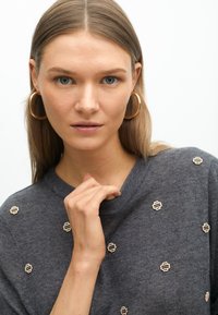 Dunkelgrauer gestrickter Pullover mit goldenen Stickereien. Das Model hat lange Haare und berührt sanft ihren Kragen, während sie direkt in die Kamera schaut.