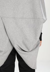 Personne portant un vêtement superposé asymétrique gris sur un pantalon noir, avec un design en tissu drapé et superposé à l'arrière.