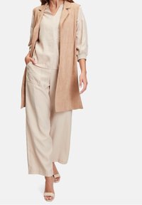 Beżowa kamizelka bez rękawów na jasnobeżowej bluzce, spodnie o szerokich nogawkach z naszytą kieszenią oraz jasne sandały na obcasie. Miękka faktura tkaniny.