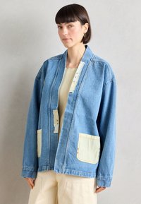 Svetlomodrá oversize džínová bunda s otvoreným predným dielom, dvoma náprsními vreckami a kontrastnými svetložltými detailmi na vreckách a golieri.