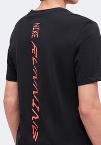 Άτομο που φοράει μαύρο μπλουζάκι Nike με κόκκινο κάθετο κείμενο "Nike Flame Running" στην πλάτη, εμφανιζόμενο από τον ώμο μέχρι τη μέση.