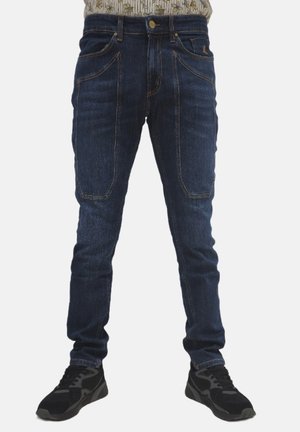Jeans in denim blu scuro con design aderente, cinque tasche e cuciture arancioni a contrasto. Indossati con scarpe da ginnastica nere.