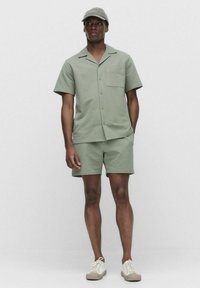 Chemise à manches courtes vert sauge avec une poche, associée à un short assorti. Texture tissée. Des baskets blanches et une casquette gris clair complètent le look.