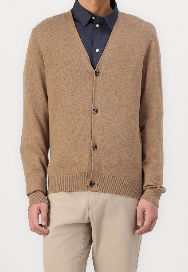 Brown knitted cardigan with a V-neck, button front, and ribbed cuffs. Worn over a dark blue collared shirt, highlighting its layered design.