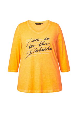 Světle oranžové tričko s výstřihem do V a třičtvrtečními rukávy, s černým nápisem „Láska je ve detailech“ na přední straně.