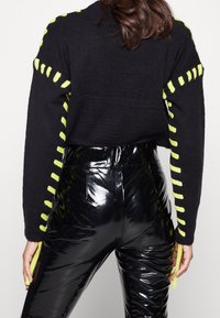 Svart stickad tröja med neongul sidodetalj, parat med glansiga svarta högmidjade byxor. Tröjan har en kortad passform och långa ärmar.