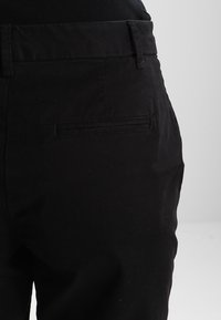 Pantalon en coton noir avec une texture lisse, doté d'une poche arrière et d'une coupe ajustée. La ceinture est visible en haut.