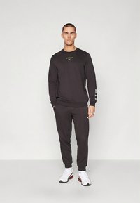 Homem em pé vestindo uma sweatshirt HYROX preta e calças de treino, com ténis brancos, contra um fundo claro simples.