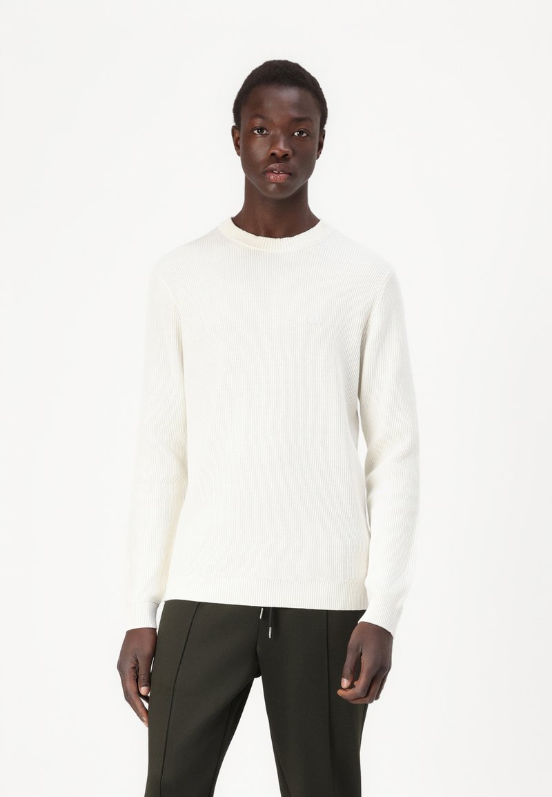 Maglione bianco a coste con scollatura a girocollo, maniche lunghe e silhouette aderente, abbinato a pantaloni verde scuro.