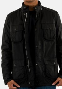 Zwarte, lichte jas met een hoge kraag, rits- en drukknoopsluiting, twee voorzakken en subtiele branding op de zak. Gelaagde stof.