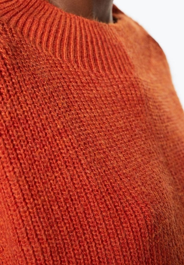 Gros plan sur une personne portant un pull orange texturé en tricot avec un col rond côtelé.