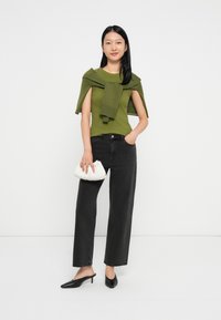 Vrouw draagt een olijfgroen top en een trui over haar schouders, zwarte brede jeans, zwarte hakschoenen en houdt een kleine witte clutch vast.