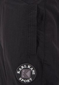 Schwarze Sporthose aus leichtem Material mit einem markanten runden Logoaufnäher mit dem Schriftzug "KARL KANI SPORT" und einer Seitentasche.