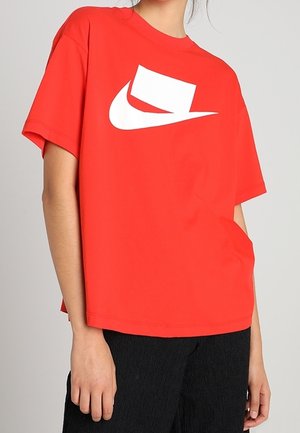 Persoon die een losse rode T-shirt draagt met een wit abstract swoosh-logo, gecombineerd met zwarte textuurbroek.