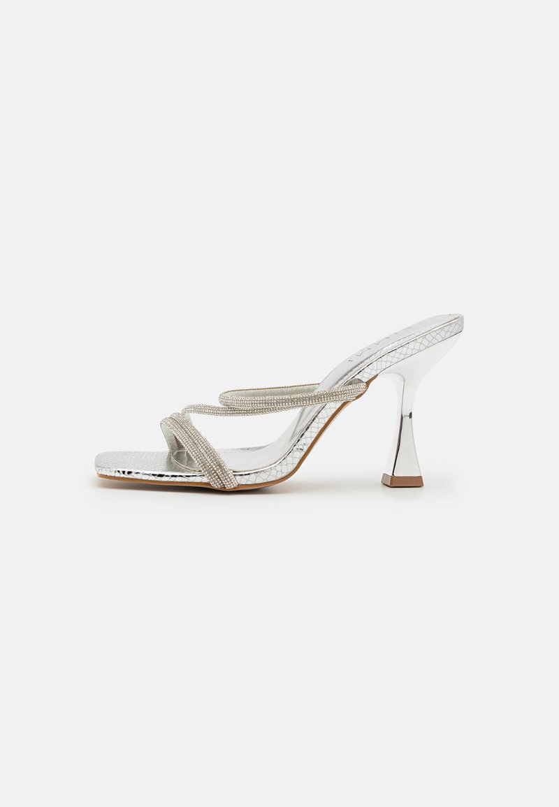 Silver heeled sandals with crisscross straps. Made of glossy material with a textured surface, featuring a squared toe and a geometric heel.