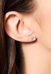 Srebrne kolczyki w kształcie serca z małymi przezroczystymi kamieniami, umiejscowione w płatku ucha, z gładkimi włosami częściowo widocznymi.