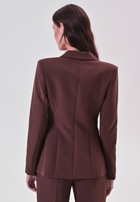 Braun maßgeschneiderter Blazer mit strukturiertem Design, ausgestattet mit schlanker Passform, Reverskragen und subtilen Nahtdetails. Glatte Stoffoberfläche.