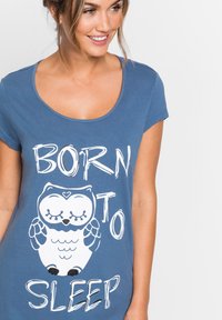 Maglietta blu a maniche corte con scollo rotondo, caratterizzata da una grafica di un gufo bianco e il testo "NATO PER DORMIRE" in carattere giocoso.