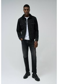 Chaqueta de mezclilla negra con un cuello de piel sobre una camiseta blanca, combinada con jeans oscuros y botas negras, presentando un corte holgado.