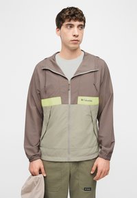 Jeune homme portant une veste Columbia bicolore et un pantalon olive, tenant un chapeau beige, regardant légèrement vers la droite avec une expression neutre.