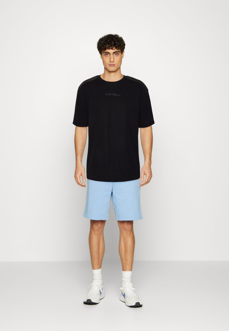Sort bomulds T-shirt med rund hals, korte ærmer og subtilt logo. Kombination med lyseblå shorts og hvide sneakers. Ensfarvet baggrund.