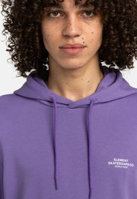 Felpa viola realizzata in morbido tessuto, con tasca frontale e cappuccio regolabile. Testo ricamato sul petto in bianco.