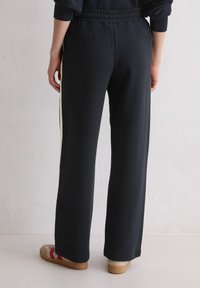 Person, der schwarze Hosen mit einem weißen Seitenstreifen und beigen Hausschuhen trägt, steht vor einer leicht strukturierten weißen Wand.