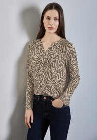 Beige Bluse mit langen Ärmeln und einem braunen abstrakten Muster, ausgestattet mit einem V-Ausschnitt und dezenten Falten, kombiniert mit dunkelblauen Jeans.
