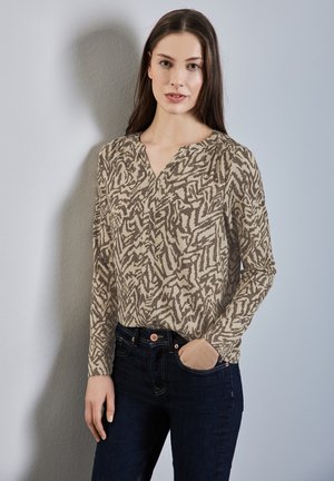 Blusa a maniche lunghe in beige con un motivo astratto marrone, caratterizzata da una scollatura a V e pieghe sottili, abbinata a jeans in denim scuro.