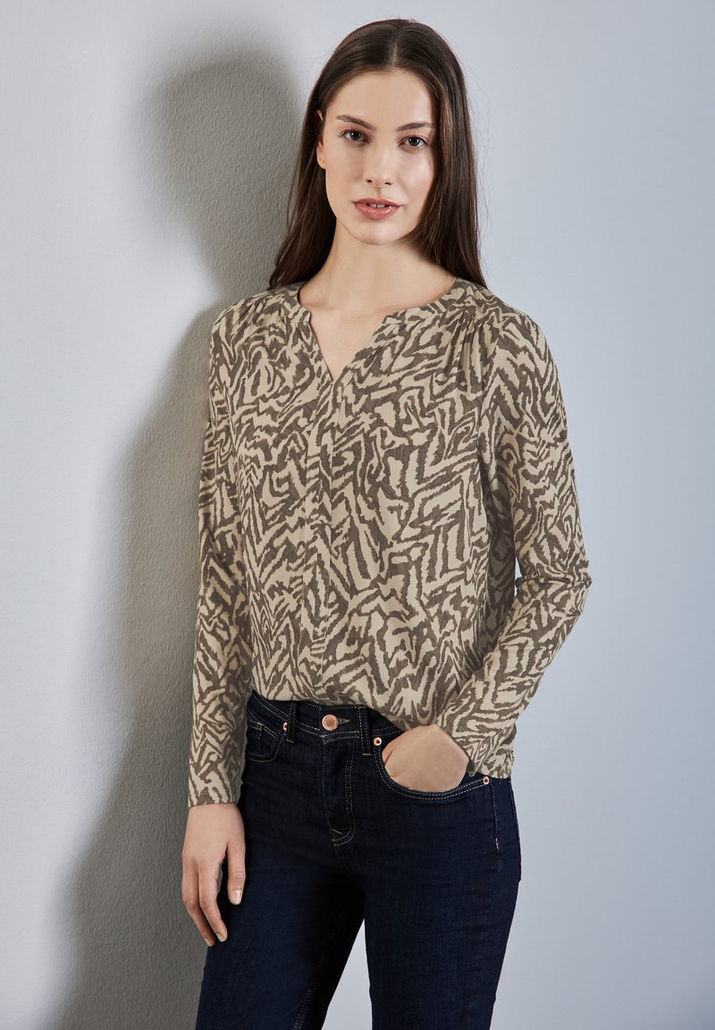Beige Bluse mit langen Ärmeln und einem braunen abstrakten Muster, ausgestattet mit einem V-Ausschnitt und dezenten Falten, kombiniert mit dunkelblauen Jeans.