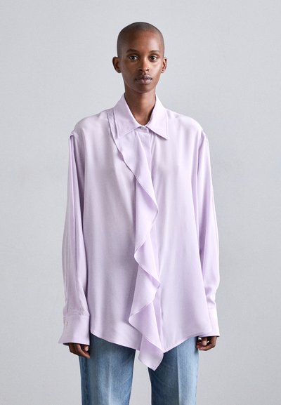 Light lavender long-sleeve blouse with a ruffled front, classic collar, and buttoned cuffs. Smooth texture, oversized fit, paired with light jeans.