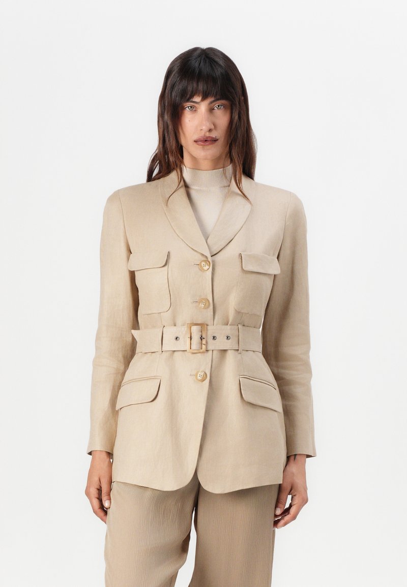Frau in beigem, tailliertem Blazer mit großen Knöpfen und Taschen, kombiniert mit passenden Hosen, die vor einem schlichten weißen Hintergrund steht.